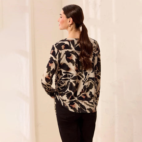 A woman shown from the back wearing a long-sleeve blouse with a black, beige, and brown abstract floral pattern, paired with black pants, set against a warm off-white background.