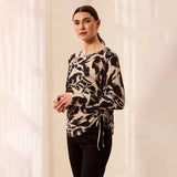 A woman modeling a long-sleeve wrap-style top with a beige, black, and brown abstract animal print, paired with black pants, standing against a softly lit cream-colored background.