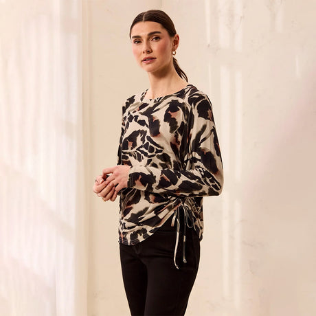 A woman modeling a long-sleeve wrap-style top with a beige, black, and brown abstract animal print, paired with black pants, standing against a softly lit cream-colored background.