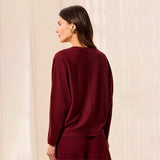 A woman shown from the back wearing a long-sleeve crewneck sweater in deep burgundy, paired with matching trousers, set against a soft beige textured background.