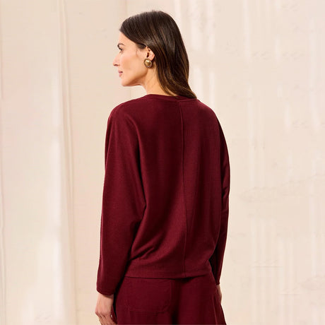 A woman shown from the back wearing a long-sleeve crewneck sweater in deep burgundy, paired with matching trousers, set against a soft beige textured background.