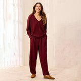 A woman modeling a matching burgundy lounge set consisting of a long-sleeve V-neck top and relaxed-fit pants, paired with tan loafers, standing in a brightly lit room with white curtains.