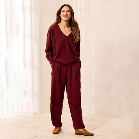 A woman modeling a matching burgundy lounge set consisting of a long-sleeve V-neck top and relaxed-fit pants, paired with tan loafers, standing in a brightly lit room with white curtains.