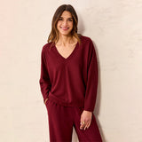 A woman wearing a matching burgundy knit loungewear set consisting of a long-sleeve V-neck top and matching pants, standing against a textured off-white wall.