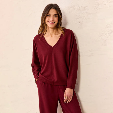 A woman wearing a matching burgundy knit loungewear set consisting of a long-sleeve V-neck top and matching pants, standing against a textured off-white wall.