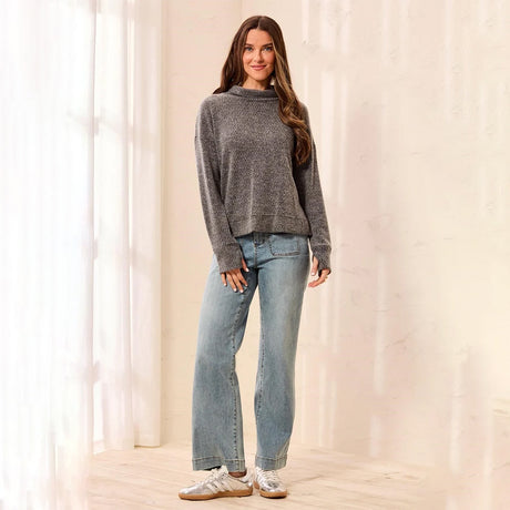 A woman stands in a softly lit room wearing a heather gray knit mock-neck sweater, light-wash blue jeans, and white sneakers.