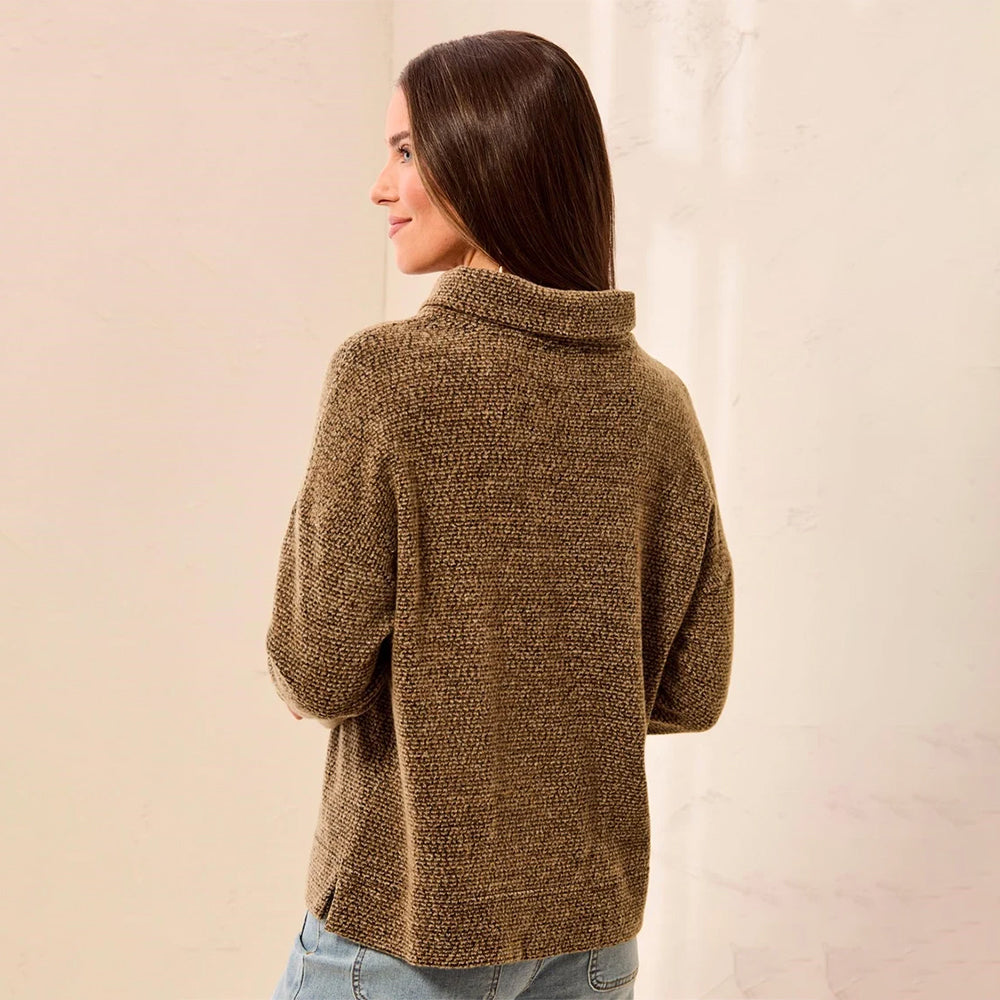 A woman with long dark brown hair is shown from the back wearing a textured, heathered brown knit turtleneck sweater and light blue jeans, standing against a light beige wall.