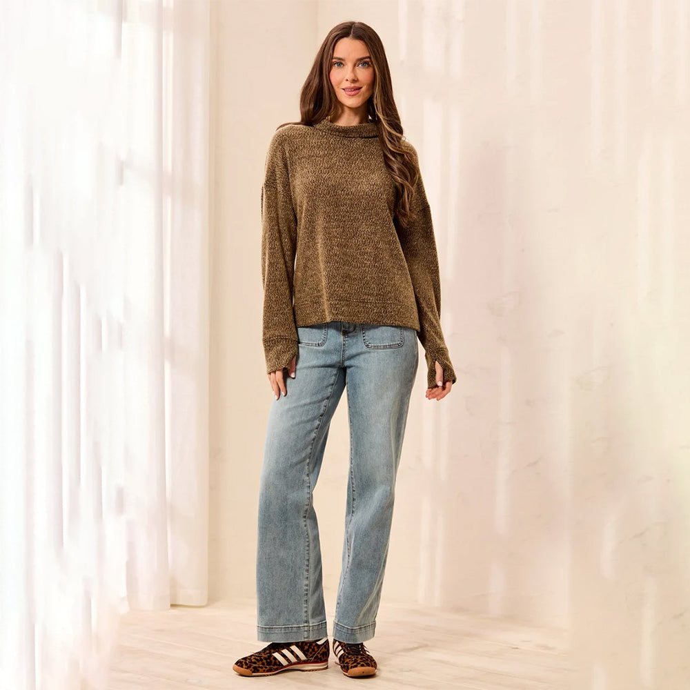 A woman stands in a bright indoor setting wearing a loose brown knit sweater, light blue straight-leg jeans, and brown-and-white striped sandals.