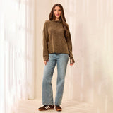 A woman stands in a bright indoor setting wearing a loose brown knit sweater, light blue straight-leg jeans, and brown-and-white striped sandals.