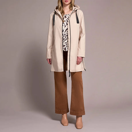 A woman models a light beige hooded raincoat over a patterned top and brown wide-leg pants, standing against a solid neutral studio background.
