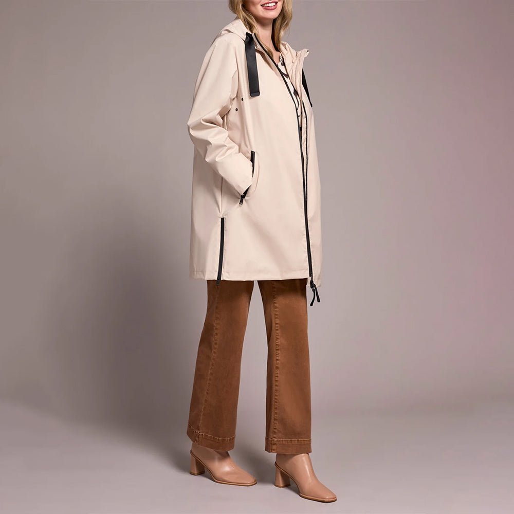A woman models a light beige hooded raincoat with black trim and zippers, paired with brown wide-leg pants and beige heels, against a solid gray studio background.