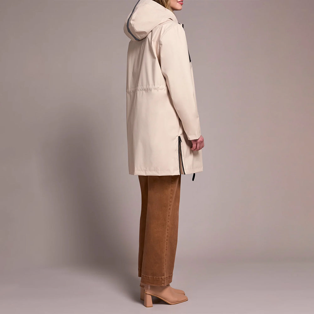 A woman shown from the side wearing a light beige hooded parka jacket, brown wide-leg pants, and tan block-heel mules against a plain warm gray studio background.
