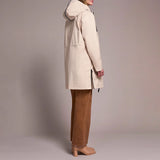 A woman shown from the side wearing a light beige hooded parka jacket, brown wide-leg pants, and tan block-heel mules against a plain warm gray studio background.