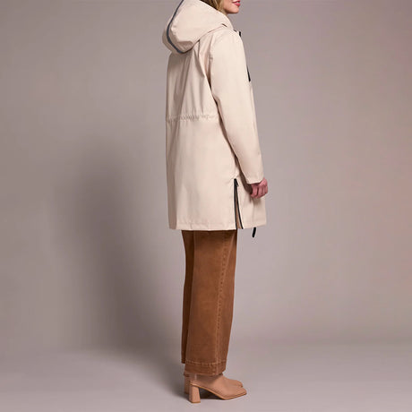 A woman shown from the side wearing a light beige hooded parka jacket, brown wide-leg pants, and tan block-heel mules against a plain warm gray studio background.