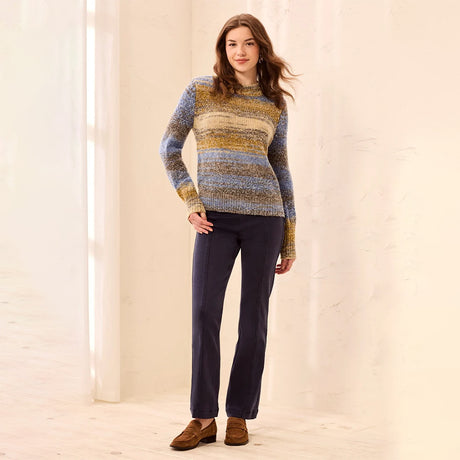 A woman stands in a bright indoor setting wearing a long-sleeve knit sweater with horizontal stripes in blue, yellow, and beige, paired with dark navy pants and brown suede loafers.