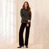 A woman modeling a black and gray leopard-print long-sleeve knit top with black wide-leg pants and dark brown heeled boots, standing in a brightly lit room with white curtains.
