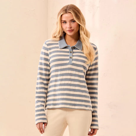 A woman modeling a long-sleeve polo shirt with horizontal gray and cream stripes, paired with light beige pants, set against a warm beige textured wall.