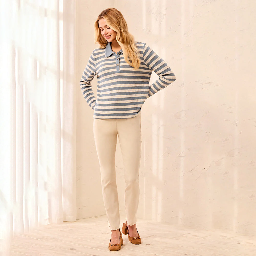 A woman modeling a long-sleeve striped polo shirt in blue and beige, paired with beige pants and brown shoes, standing in a bright, sunlit room with sheer curtains.