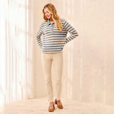 A woman modeling a long-sleeve striped polo shirt in blue and beige, paired with beige pants and brown shoes, standing in a bright, sunlit room with sheer curtains.