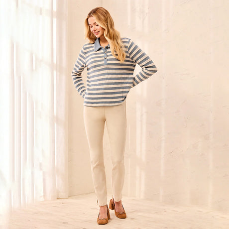 A woman modeling a long-sleeve striped polo shirt in blue and beige, paired with beige pants and brown shoes, standing in a bright, sunlit room with sheer curtains.