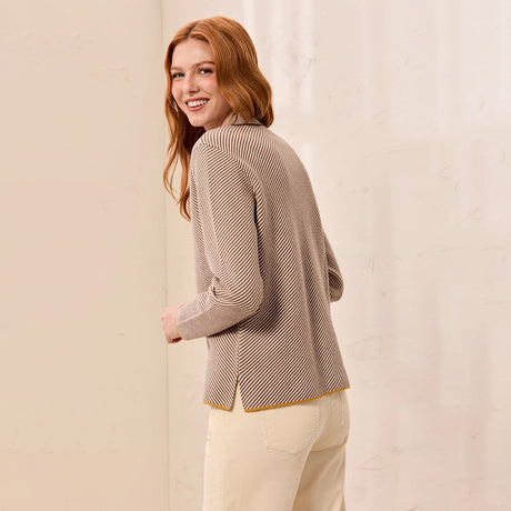 A woman with red hair is shown from the back wearing a beige and brown striped knit sweater and cream pants, standing against a light beige wall.