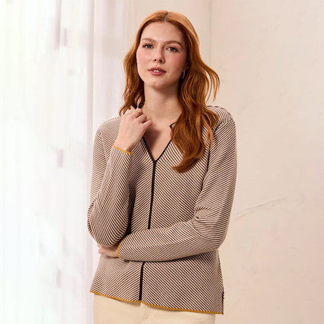 A woman with red hair wearing a beige and black striped long-sleeve blouse with gold trim, standing against a light cream background.