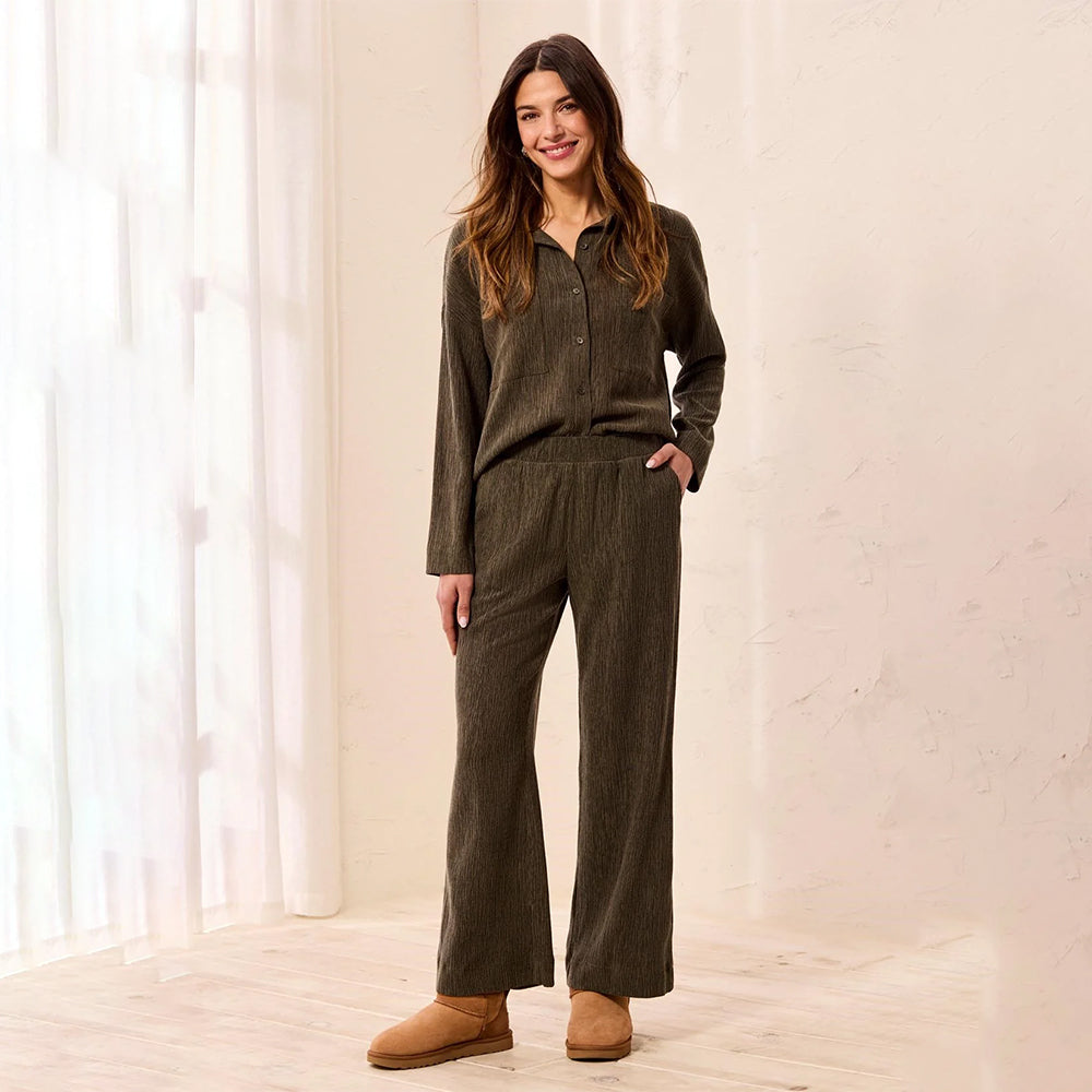 A woman modeling a matching two-piece lounge set in a warm, softly lit room. The set consists of a long-sleeve button-front top and matching wide-leg pants, both in a textured olive-brown fabric.