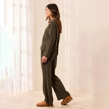 A woman wearing a matching dark olive green lounge set consisting of a cropped long-sleeve top and wide-leg pants, paired with brown suede slippers, standing in a brightly lit room with sheer curtains.