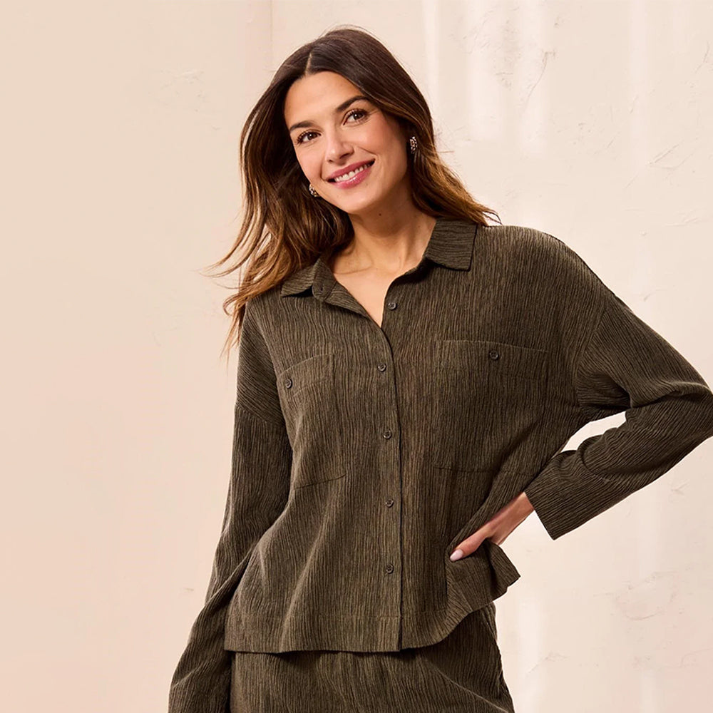 A woman wearing a dark olive green corduroy button-up shirt and matching pants, posing against a light beige textured wall.