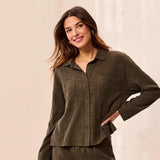 A woman wearing a dark olive green corduroy button-up shirt and matching pants, posing against a light beige textured wall.