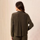 A woman shown from the back wearing a textured, dark olive green long-sleeve top and matching pants against a neutral cream-colored wall.