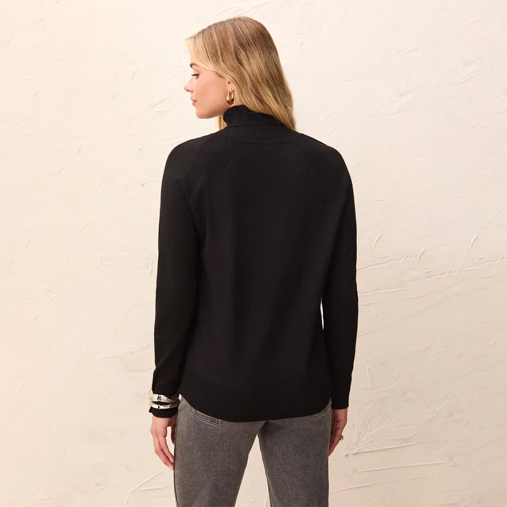 A woman shown from the back wearing a black long-sleeve mock neck sweater and gray pants, standing against a textured off-white wall.