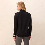 A woman shown from the back wearing a black long-sleeve mock neck sweater and gray pants, standing against a textured off-white wall.