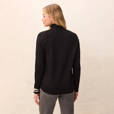 A woman shown from the back wearing a black long-sleeve mock neck sweater and gray pants, standing against a textured off-white wall.