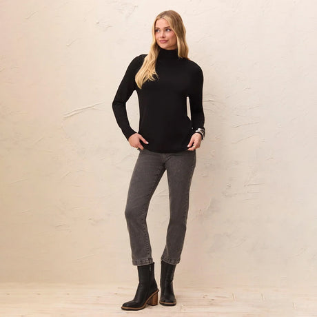 A woman modeling a black long-sleeve mock neck turtleneck sweater, paired with dark gray jeans and black ankle boots, standing against a textured beige wall.