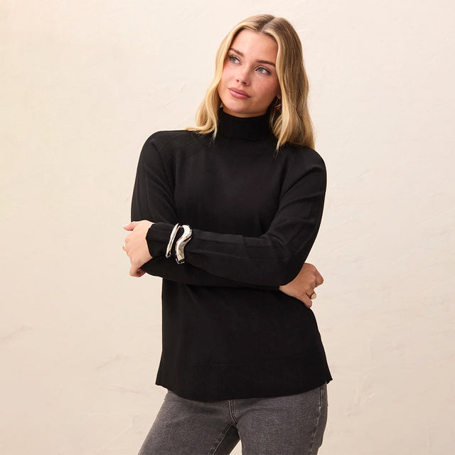 A woman wearing a black ribbed long-sleeve mock neck sweater with white cuff stripes, paired with gray jeans, standing against a light beige textured wall.