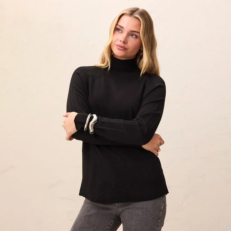 A woman wearing a black ribbed long-sleeve mock neck sweater with white cuff stripes, paired with gray jeans, standing against a light beige textured wall.
