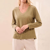 A woman wearing a long-sleeve V-neck top in a muted olive green color, paired with light beige pants, against a soft beige background.