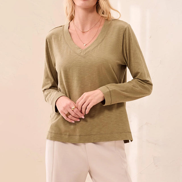 A woman wearing a long-sleeve V-neck top in a muted olive green color, paired with light beige pants, against a soft beige background.