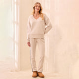 A woman modeling a matching beige lounge set consisting of a V-neck sweatshirt and straight-leg sweatpants with thin dark side stripes, paired with tan boots, in a brightly lit cream-colored room.