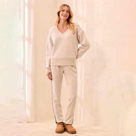 A woman modeling a matching beige lounge set consisting of a V-neck sweatshirt and straight-leg sweatpants with thin dark side stripes, paired with tan boots, in a brightly lit cream-colored room.