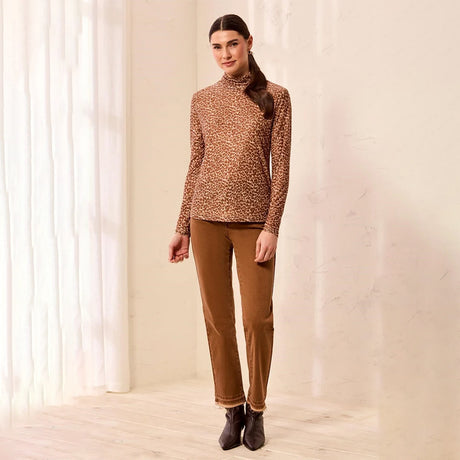 A woman modeling a brown leopard-print mock-neck long-sleeve knit top with brown pants and dark ankle boots, standing in a brightly lit room with white curtains and a light wall.
