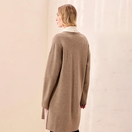 A woman shown from the back wearing an oversized taupe knit cardigan with a contrasting cream collar, standing against a textured off-white wall.