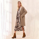 A woman models a long taupe knit cardigan over a brown and beige patterned midi dress, paired with brown ankle boots, standing in front of a white curtain backdrop.