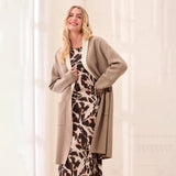 A woman modeling a long taupe cardigan over a brown and beige animal print dress, standing in a bright, neutral-toned indoor setting.