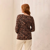 A woman with red hair is shown from behind wearing a brown and black leopard-print long-sleeve top and cream pants, standing against a light textured wall.