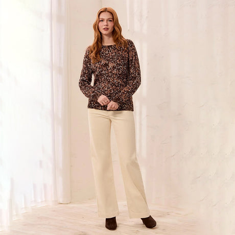 A woman stands in a bright indoor setting wearing a long-sleeve knit top with a dark brown and black abstract pattern, paired with cream-colored wide-leg pants and dark brown shoes.