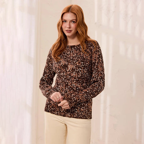 A woman with red hair wearing a brown leopard-print long-sleeve top and cream pants, standing against a softly lit cream-colored wall.