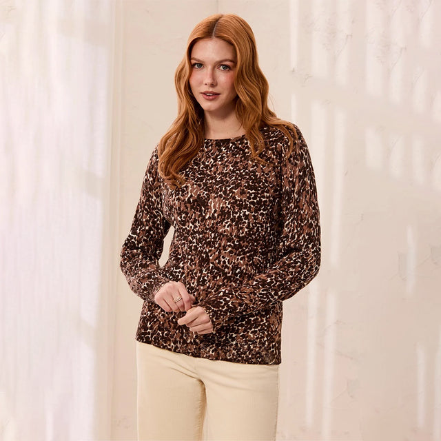 A woman with red hair wearing a brown leopard-print long-sleeve top and cream pants, standing against a softly lit cream-colored wall.