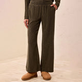 A person wearing dark olive green wide-leg knit pants with a matching long-sleeve top and tan suede boots, standing against a light beige wall and tiled floor.